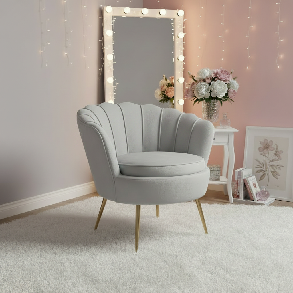 A light gray upholstered accent chair with gold-tone legs and two matching gray floral throw pillows, styled in a softly lit room with a mirror, string lights, flowers, and a small side table.