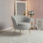 A light gray upholstered accent chair with gold-tone legs and two matching gray floral throw pillows, styled in a softly lit room with a mirror, string lights, flowers, and a small side table.