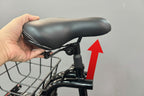 A close-up of a black bicycle saddle being held by a hand, showing the textured leather-like surface and white contrast stitching. The saddle is mounted on a black bicycle frame with a front basket, set against a blurred indoor background.