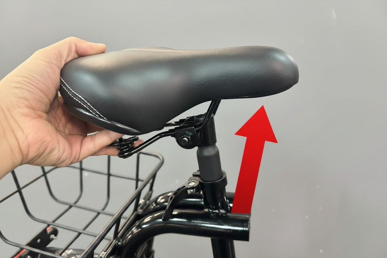 A close-up of a black bicycle saddle being held by a hand, showing the textured leather-like surface and white contrast stitching. The saddle is mounted on a black bicycle frame with a front basket, set against a blurred indoor background.