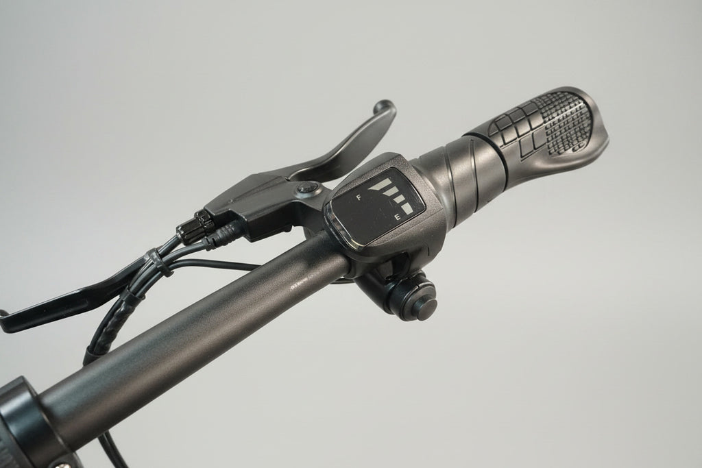 Close-up of a black electric bicycle handlebar with a digital throttle control display, showing the throttle grip and brake lever in a workshop setting.