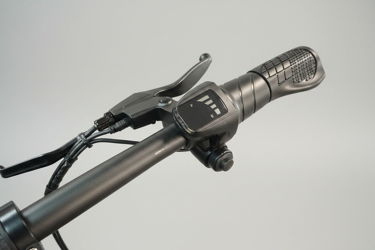 Close-up of a black electric bicycle handlebar with a digital throttle control display, showing the throttle grip and brake lever in a workshop setting.