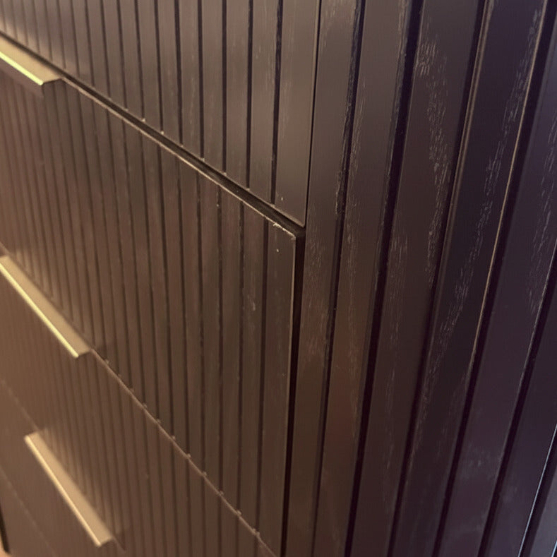 Close-up of a dark brown wooden cabinet or dresser with vertical ribbed drawer fronts and small rectangular metal pulls, photographed indoors.