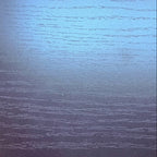 A close-up of a dark wood grain surface, likely a table top, with a blue reflective sheen and a person's shoes visible at the bottom edge.