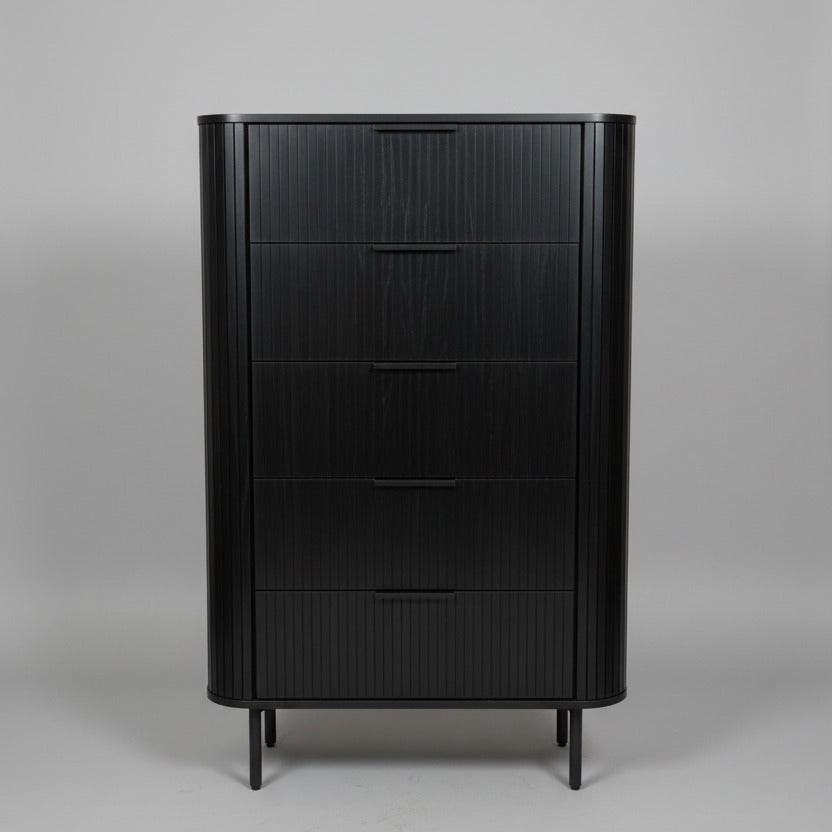 A tall black wooden chest of drawers with four drawers and a ribbed front, standing on small legs in a showroom or office next to a window.
