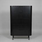 A tall black wooden chest of drawers with four drawers and a ribbed front, standing on small legs in a showroom or office next to a window.