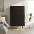 A tall black ribbed dresser or chest with five drawers stands in a modern, sunlit bedroom next to a bed and a small potted plant.