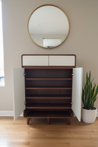 Wooden shoe cabinet with open doors on a wooden floor, wall mirror above, and plant to the side.