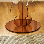 A modern round side table with a clear glass top and a sculptural amber-brown mirrored pedestal base, styled in a living room next to a dark leather armchair.