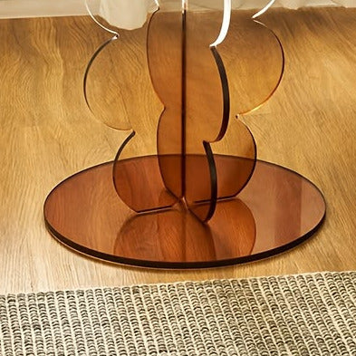 A modern round side table with a clear glass top and a sculptural amber-brown mirrored pedestal base, styled in a living room next to a dark leather armchair.
