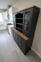A large black wooden hutch and wine cabinet with glass-front upper doors, open shelving, a wine rack, and two wood-look countertops, shown in a room with white walls and a dark floor.