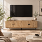A light natural wood TV stand or media console with black legs and round black knobs, shown in a styled living room setting below a wall-mounted flat-screen TV. The room features a neutral beige wall, a woven rug, and decorative plants and vases.